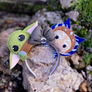 May include: A brown headband with a brown bow and two felt character ears. The left ear is green and pink and features a character with large eyes. The right ear is blue and brown and features a character with a white face and blue and brown stripes.