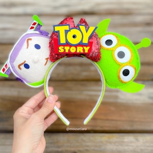 May include: Toy Story themed Minnie Mouse ears headband with a red bow, a green alien character, and a purple character with the words "Toy Story" on the bow.