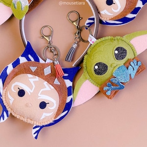 May include: Three felt keychains featuring characters from popular movies. The first keychain is a blue and white character with a brown face and white markings. The second keychain is a green character with a frog on its chest. The third keychain is a brown and white character with a striped pattern. The keychains are attached to silver key rings.
