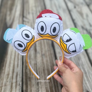 Puede incluir: Una diadema amarilla con tres personajes de pato de fieltro blanco unidos. Los patos tienen ojos negros, picos naranjas y sombreros rojos, azules y verdes.