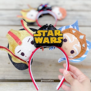 May include: A headband featuring two circular ears decorated with felt character faces, one in gold and red and the other in blue and white. A black sequined center piece displays the yellow glitter text STAR WARS. A second headband is visible in the background.