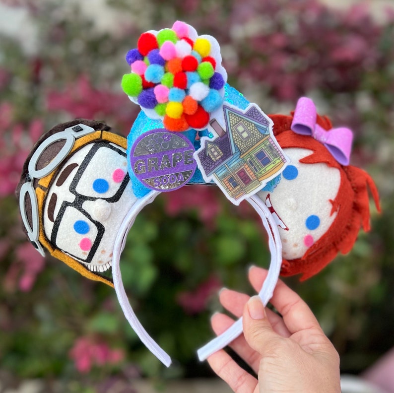 May include: Up themed Minnie Mouse ears with a felt character of Carl Fredricksen and Russell, a house, a bunch of balloons, and a Grape Soda pin.