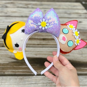 Può includere: Un cerchietto bianco con un fiocco di paillettes viola, un personaggio di feltro giallo con capelli neri e un viso bianco, e un personaggio di feltro marrone con orecchie rosa e occhi blu. Il cerchietto è decorato con fiori di feltro bianchi.