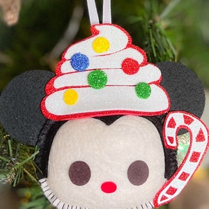May include: A felt Christmas ornament featuring a cartoon character's face with black ears, a white face, and a red mouth. The ornament is topped with a white, red, and glittery frosting design with colorful dots and a candy cane.