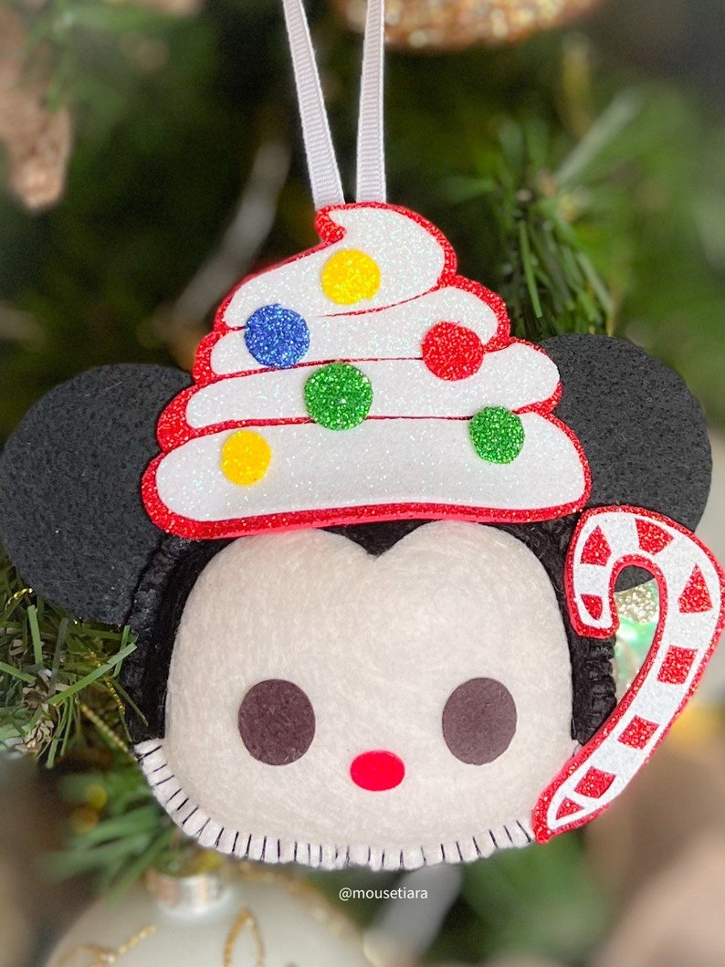 May include: A felt Christmas ornament featuring a cartoon character's face with black ears, a white face, and a red mouth. The ornament is topped with a white, red, and glittery frosting design with colorful dots and a candy cane.