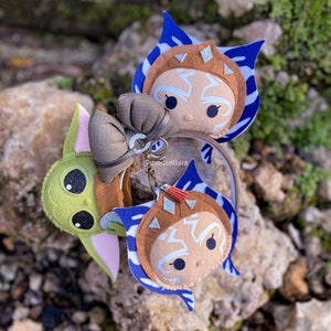 May include: A headband with two felt character charms. One charm is a green Baby Yoda character and the other is a brown Ahsoka Tano character. The headband is brown with a brown bow.