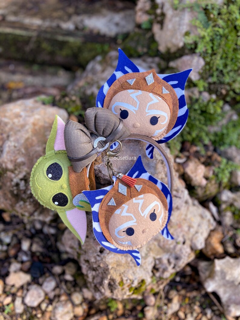 May include: A headband with two felt character charms. One charm is a green Baby Yoda character and the other is a brown Ahsoka Tano character. The headband is brown with a brown bow.