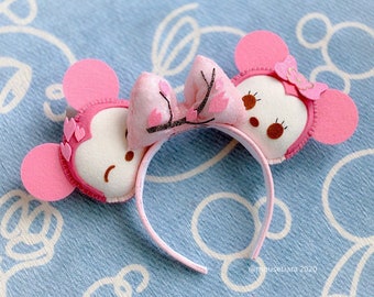 May include: Pink Minnie Mouse ears with a pink and white bow and a cherry blossom design. The ears are made of felt and have a white headband.