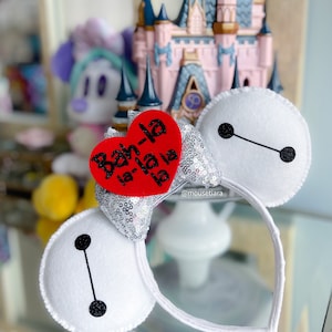 May include: White felt headband with a red heart that says "Bah-la la-la la" and a silver sequin bow. The headband is shaped like the character Baymax from the Disney movie Big Hero 6.