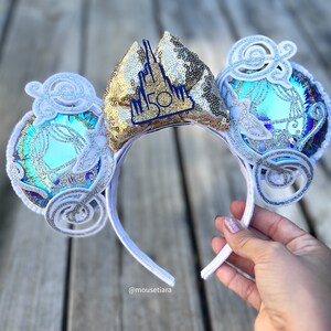 May include: White Minnie Mouse ears with a gold sequin bow featuring a blue castle with the number 50 on it. The ears are decorated with white swirls and iridescent blue and silver glitter.