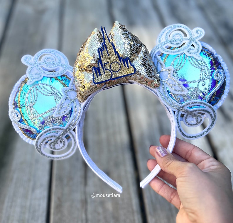 May include: White Minnie Mouse ears with a gold sequin bow featuring a blue castle with the number 50 on it. The ears are decorated with white swirls and iridescent blue and silver glitter.