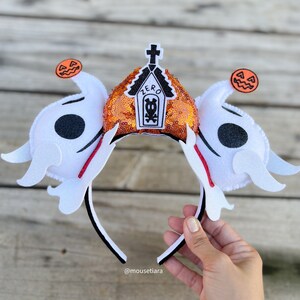May include: Orange and white Halloween headband with a black and white tombstone with the word "ZERO" on it. The headband has two white ghost ears with black eyes and orange pumpkin accents.