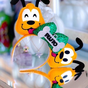 Peut inclure: Un serre-t&ecirc;te en feutre jaune et noir avec un personnage en peluche de Pluto portant un n&oelig;ud vert et argent&eacute; paillet&eacute; avec un os sur lequel est &eacute;crit "Pluto".