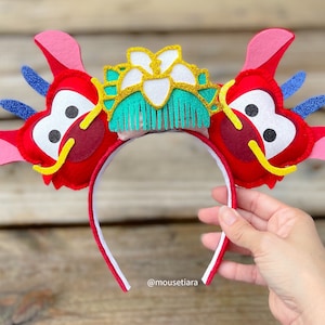 May include: Red and pink felt mouse ears with a gold and green felt dragon head and a white felt flower on top. The ears are attached to a white headband with a red trim. The headband is held by a hand. The text "@mousetiara" is visible in the bottom right corner of the image.