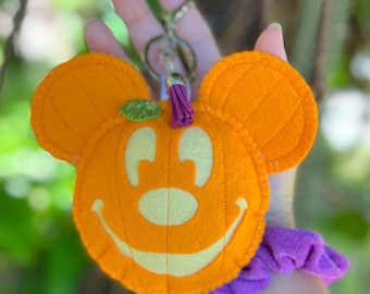May include: Orange felt Mickey Mouse pumpkin keychain with a smiling face, green glitter leaf, and purple tassel. The keychain is attached to a silver ring. The background is blurred green.