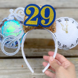 May include: White headband with decorative ears. One ear features a clock face with gold hands and Roman numerals. The other ear has a blue and gold number 29. A third ear has a blue and iridescent design with silver accents. The headband is white.