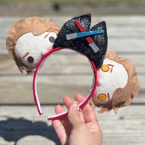 May include: A pair of red and white mouse ears with a black and silver sequin ear featuring a red and blue lightsaber design. The ears are attached to a red headband.