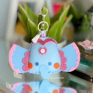 Puede incluir: Un llavero de elefante de fieltro azul con detalles en rosa y blanco. El elefante tiene un borla rosa y un llavero plateado. El llavero está decorado con purpurina.
