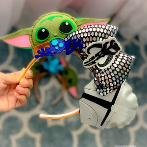 May include: A pair of handmade Disney-themed Minnie Mouse ears. One ear features a green and pink felt Baby Yoda character. The other ear features a silver sequin bow with a black and white Star Wars character.
