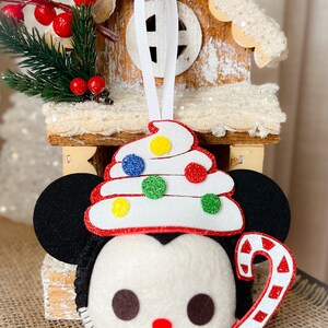 May include: A felt Christmas ornament featuring a cartoon character's face with black ears, a white face, and a red nose. The ornament has a white, red-trimmed hat decorated with colorful glitter dots and a candy cane. A white ribbon is attached for hanging.