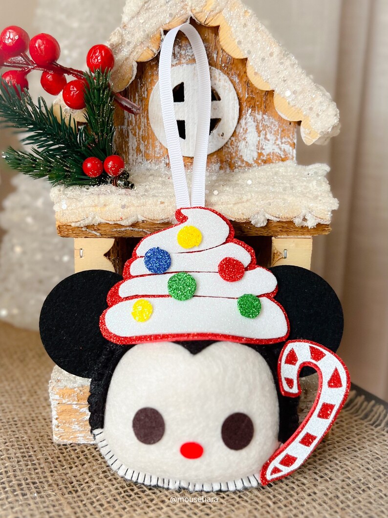 May include: A felt Christmas ornament featuring a cartoon character's face with black ears, a white face, and a red nose. The ornament has a white, red-trimmed hat decorated with colorful glitter dots and a candy cane. A white ribbon is attached for hanging.