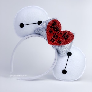 May include: White felt headband with two white felt Baymax ears. Each ear has a black circle and a black line. A red heart with the text "Baymax" in black is attached to a silver sequin bow.