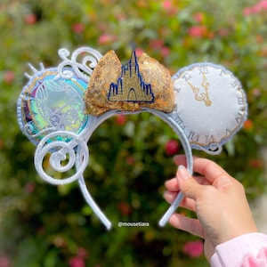 May include: A silver headband with three decorative elements. One ear features a gold sequined castle, another a clock face, and the third a circular design with a holographic effect. The headband is held by a hand, with a pink scrunchie visible.