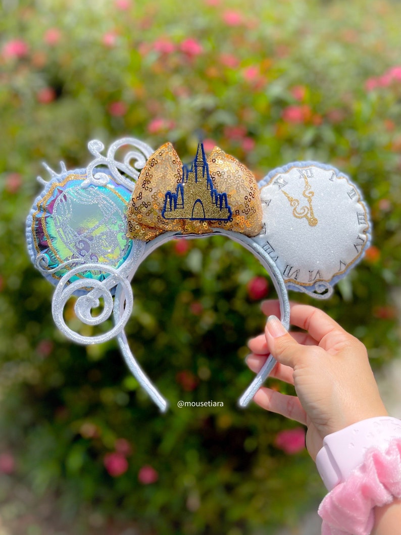May include: A silver headband with three decorative elements. One ear features a gold sequined castle, another a clock face, and the third a circular design with a holographic effect. The headband is held by a hand, with a pink scrunchie visible.