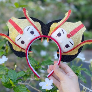 May include: A pair of black mouse ears with gold and red glitter fabric. The ears feature a cartoon face with a crown and a red and gold design.