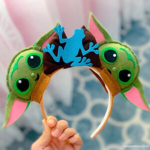 May include: Green felt headband with a pair of ears and a blue felt frog silhouette. The ears are pink on the inside and have a brown felt trim.