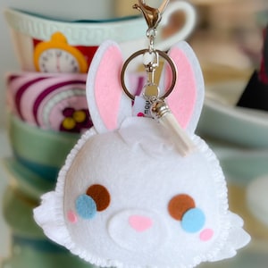 Può includere: Portachiavi a forma di coniglio in feltro bianco con orecchie rosa, occhi marroni e blu e una coda in feltro bianco. Il portachiavi ha un anello in metallo argentato e una chiusura in metallo argentato.