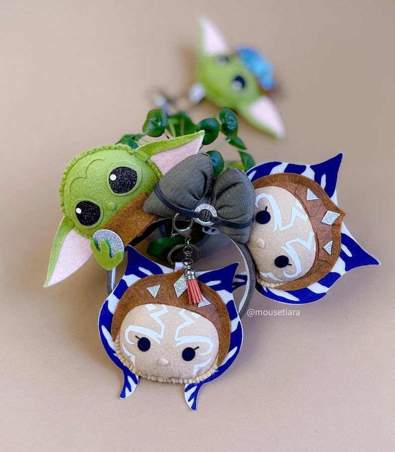 May include: Three felt character keychains: a green Baby Yoda, a brown Ahsoka Tano, and a brown Ahsoka Tano with blue and white stripes. The keychains are attached to a silver key ring with a brown tassel.