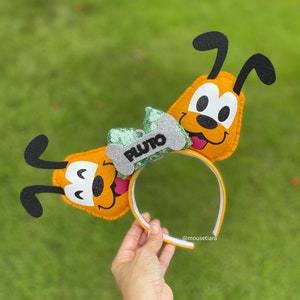 Peut inclure: Une paire d'oreilles de Pluto en feutre jaune avec un n&oelig;ud vert et argent et un os sur lequel est inscrit "PLUTO". Les oreilles sont attach&eacute;es &agrave; un bandeau noir.