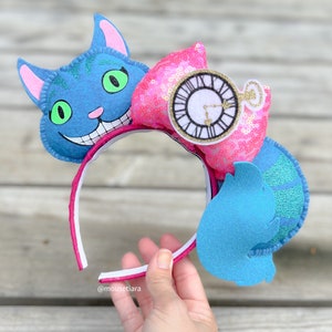 May include: Pink and blue felt headband with a Cheshire Cat, a pocket watch, and a blue and green character from Alice in Wonderland.