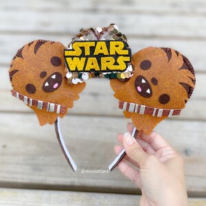 May include: Brown felt mouse ears with a Chewbacca character design. The ears have a gold sequin bow with the text "STAR WARS" in black letters.