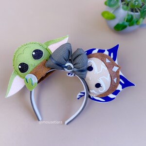 May include: A pair of felt character ears for a headband. The left ear is green and features a character from Star Wars: The Mandalorian. The right ear is blue and white and features a character from Star Wars: The Clone Wars. The headband is black and white.