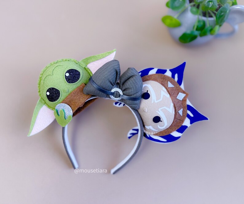 May include: A pair of felt character ears for a headband. The left ear is green and features a character from Star Wars: The Mandalorian. The right ear is blue and white and features a character from Star Wars: The Clone Wars. The headband is black and white.