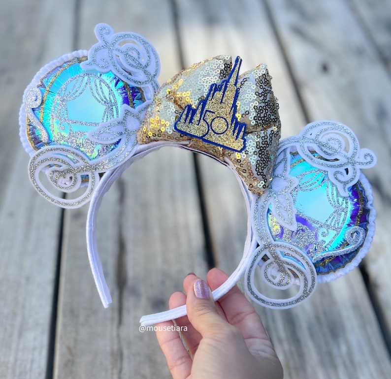 May include: White headband with a gold and blue bow featuring a Cinderella carriage and the text "50th" on the bow.