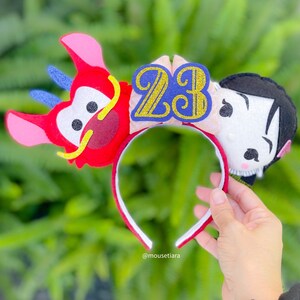 May include: Red and white felt Mickey Mouse ears with a gold and blue number 23 on the headband. The ears are shaped like Mulan and Mushu from the Disney movie Mulan.