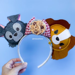 May include: A pair of felt mouse ears featuring a grey and white dog, a brown and white dog, and a plate of spaghetti with meatballs. The ears are attached to a white headband. The headband is held by a hand. The text '@mousetiara' is visible on the headband.