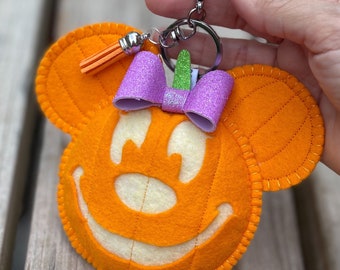 Pode incluir: Chaveiro de feltro laranja em forma de abóbora com um laço de purpurina roxo e um pequeno tassel laranja. O chaveiro está preso a um anel de chave prateado.