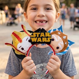 May include: A headband featuring two circular ears with character designs and a central black plaque with the yellow text STAR WARS. The left ear is gold and white with red accents, while the right ear is blue and white with tan details.