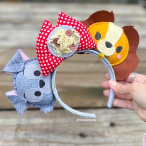 May include: A pair of gray, brown, and red felt mouse ears with a white headband. The ears are decorated with a plate of spaghetti and meatballs, a cartoon dog face, and a cartoon wolf face. The headband has the text "@mousetiara" printed on it.