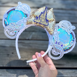 May include: White headband with a blue and silver Cinderella carriage design on each ear. The headband also features a gold bow with a blue castle and "50" on it.