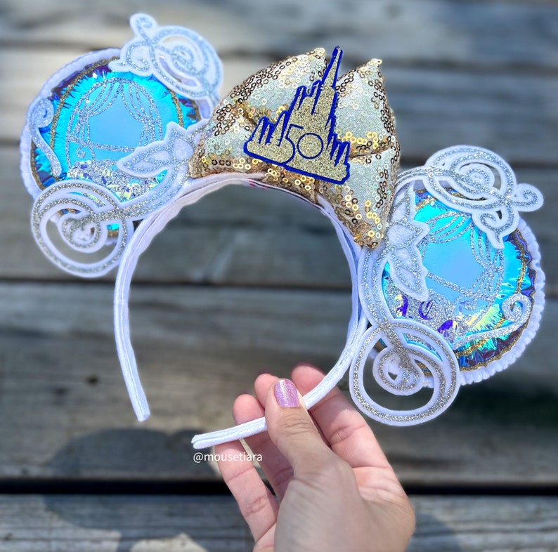 May include: White headband with a blue and silver Cinderella carriage design on each ear. The headband also features a gold bow with a blue castle and "50" on it.