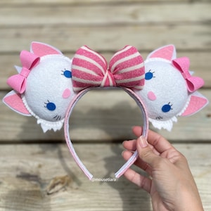 Può includere: Fascia per capelli con orecchie di gatto in feltro bianco, fiocco a righe rosa e bianco, occhi blu e dettagli glitter rosa. La fascia è realizzata in feltro bianco e ha un fiocco a righe rosa e bianco sulla parte superiore. Le orecchie sono a forma di orecchie di gatto e hanno occhi blu e dettagli glitter rosa.