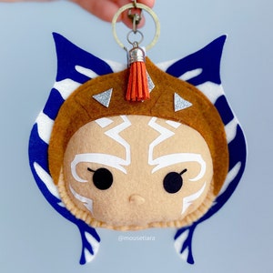 May include: A felt keychain featuring a cartoon-style Ahsoka Tano from Star Wars. The keychain is brown, blue, and white with a silver key ring and an orange tassel.