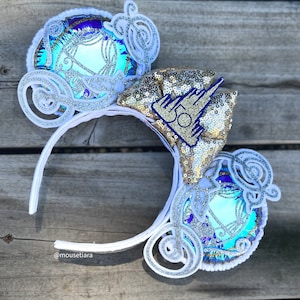 May include: White mouse ears with a blue and gold Cinderella carriage design. The bow on the ears features gold sequins and a blue castle with the number 50 on it.