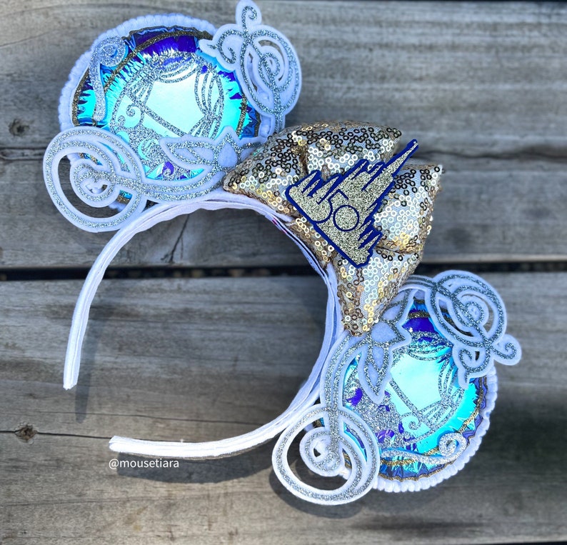 May include: White mouse ears with a blue and gold Cinderella carriage design. The bow on the ears features gold sequins and a blue castle with the number 50 on it.