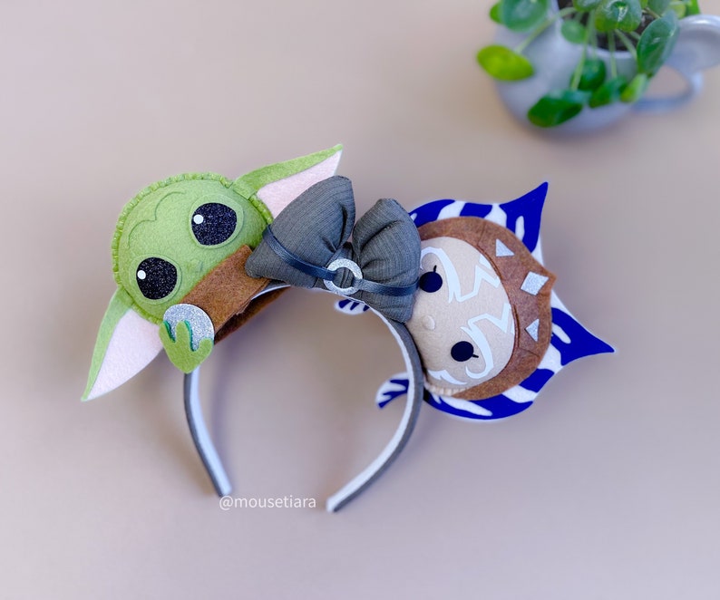 May include: A pair of felt character headbands featuring a green Baby Yoda and a blue and brown Ahsoka Tano. The headbands are attached to a black headband with a gray bow.